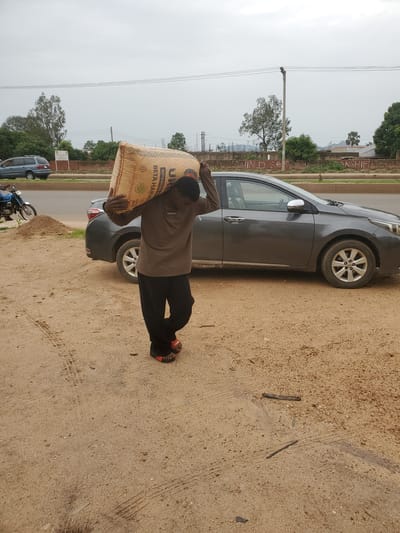 A man carries a large sack of grain over his shoulder, walking on a dusty path. He is wearing a brown long-sleeved shirt and black pants. He has red sandals on his feet. A gray sedan is parked behind him on a paved road. In the background, there are trees, a blue minivan, and utility poles. The sky is overcast. The scene is in an urban area in Unknown, Nigeria.