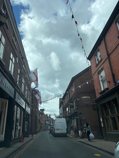 Here is a description of the image:
The image shows a street in Knutsford, United Kingdom, with a slightly tilted perspective. Buildings line both sides of the street, primarily constructed of red brick. Festive bunting and English flags are strung across the street, adding a celebratory atmosphere. A white van is parked on the street. People are seen walking on the sidewalk. Shop signs indicate businesses such as "Clarence Paul" and "Margao" restaurant. The sky is cloudy. Double yellow lines are marked on the road. The photograph appears to be taken from a low angle within the street itself.