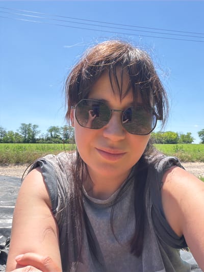 In Ust'-Dzhigutinka, Russia, on a bright, sunny day, a woman with dark hair and bangs wears large, hexagonal sunglasses and a gray t-shirt. She is seated outdoors, possibly in a field, with a background of green grass, trees, and a clear blue sky. Power lines are visible against the sky. The sunglasses reflect a cell phone in her hand and the surrounding environment. Her left arm and shoulder are visible, with a portion of her left hand in the foreground.