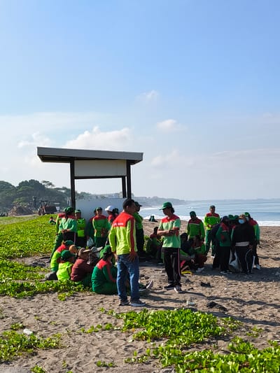 A group of approximately 25-30 people are gathered on a beach in Kerobokan Kelod, Indonesia. They are wearing green and red shirts, and some have green hats. Some are standing, while others are sitting on the sand. They are positioned near a white sign structure. The backdrop features a beach with ocean waves and some vegetation. The sky is blue with scattered clouds. The ground is covered with sand and some patches of green plants. There is a tractor parked on the grassy section of the beach, in the background, to the left. The scene appears to be a gathering or break during some activity, potentially a community service event or beach cleanup.