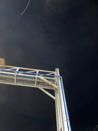 The image shows a nighttime view looking up at a metal structure, possibly part of an overpass or a support for utilities, against a dark sky. The structure is white with some rust showing and has blue pipes running along it. In the upper left quadrant of the sky, there is a faint white streak, possibly a contrail from an aircraft or a shooting star. A single small bright point, likely a star or planet, is visible in the upper right quadrant of the sky. The background is a deep, dark blue, indicating it is nighttime. The scene appears to be in an outdoor urban or semi-urban environment. There are no people or animals visible. The lighting is dim, suggesting artificial light sources are not prominently illuminating the sky.
