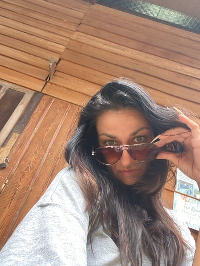 In Thôn 3, Vietnam, a woman with long, dark hair and green eyes is taking a selfie. She is wearing a grey sweatshirt and sunglasses. Her right hand is holding the sunglasses up, revealing her eyes. The background is made of wooden planks. There is a sign with Vietnamese text and a picture of what appears to be a resort or eco-lodge.