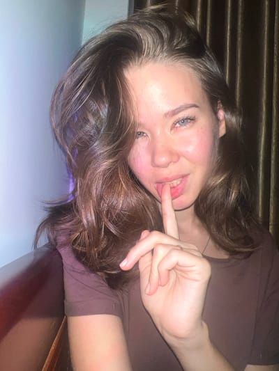 A young woman with long, wavy, dark brown hair is captured in a close-up, horizontal portrait, looking directly at the camera. Her head is tilted, and she appears to be lying down on her side. She has light blue or grey eyes, a soft smile, and her left index finger is gently placed on her lower lip, suggesting a playful or thoughtful expression. She has a natural blush on her cheeks and her lips are glossy. She is wearing a simple dark brown or maroon colored top.
The background behind her head is dark and features horizontal textures, possibly blinds or wooden paneling. Her hair spills over a light blue-grey surface in the foreground, likely a bed or table. To the right, a darker, reddish-brown wooden surface is visible. The lighting is dim, casting a soft, warm glow, indicating an indoor setting, possibly in the evening or at night. There are no discernible location cues within the image that specifically point to Nam Natural Heritage Area Trang, Vietnam; it depicts a generic interior. No text is visible in the image.