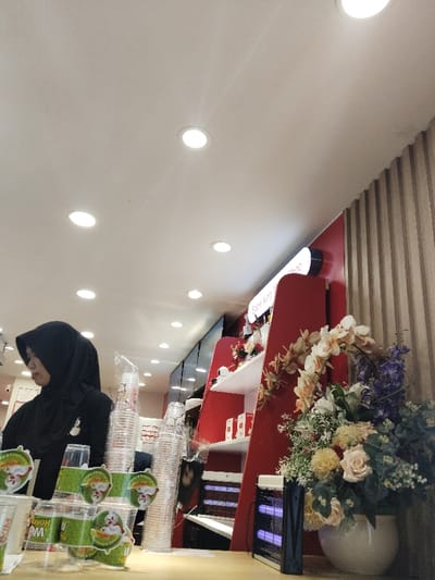 The image shows a woman in a black hijab and black clothing standing behind a counter in a store. The woman is looking down at a stack of plastic cups in front of her. To her right, there is a red and white display shelf, with a large flower arrangement in a white pot, a black lamp with a purple light, and stacks of plastic cups on the counter below it. The ceiling has multiple recessed lighting fixtures. The store is located at the coordinates -6.338, 107.166 in Bekasi Regency, Indonesia. The environment is well-lit and appears to be a retail store.