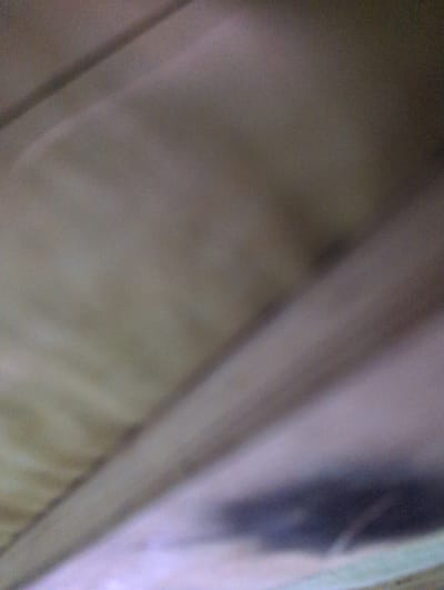 The image presents an abstract, heavily blurred, and out-of-focus scene, attributed to the city of Chaguanas, Trinidad and Tobago. The composition consists primarily of muted tones, ranging from light beige-gray to darker purplish-gray and brown. Diagonal lines or folds traverse the frame, originating from the upper left and extending downwards towards the right, creating a sense of layered or textured surfaces. A distinct, amorphous dark gray shape occupies the lower central-right portion of the frame, possibly representing a shadow or a deeply shaded area. The lighting is diffuse, with subtle gradations between lighter and darker regions across the visual field. There are no discernible people, specific objects, or overt actions or interactions depicted. The overall environment appears to be an indistinct, close-up view of a surface, possibly fabric or a wall, under low or uneven illumination.