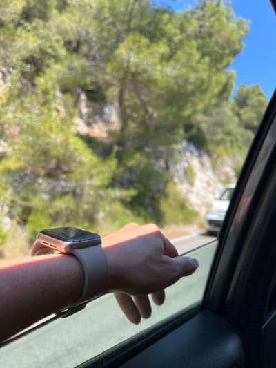 An arm with a smartwatch rests on a car window, with the hand slightly extended outside. The background shows a sunlit coastal road with green foliage and a blue sky. The scene is likely near Mattinata, Italy, a town known for its scenic coastal roads.