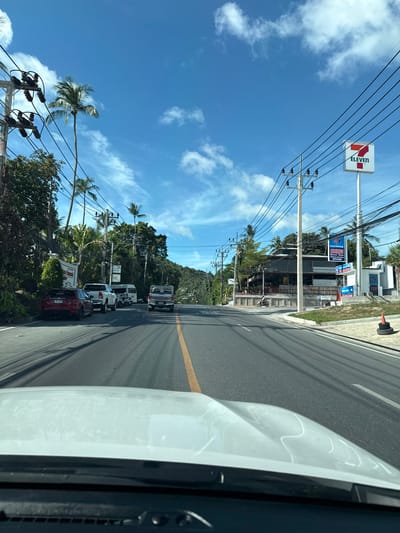 The image depicts a street scene in Baan Chaweng Noi, Thailand, on a clear day with blue skies and scattered clouds. The perspective is from the driver's seat of a vehicle, looking forward down a paved road. On the left side of the road, there are dense green trees and palm trees, a sign that reads "Samui Summit," and several parked vehicles including a red sedan, a white pickup truck, and a white van. Further down the road, a white Toyota pickup truck is visible.
On the right side of the road, a prominent 7-Eleven convenience store sign is visible. Adjacent to it is a building with a roof extending over a dining area, with tables and chairs. A currency exchange sign with blue lettering is also present. A few motorcycles are parked near the buildings, and a person is seen riding a motorcycle. Electrical poles with numerous wires run along both sides of the street. The road is marked with a yellow center line and has shadows cast by overhead wires. A traffic cone made of stacked tires is present on the right shoulder of the road.