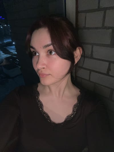 A young woman with auburn hair, pale skin, and brown eyes is pictured from a close-up, angled perspective. She wears a dark top with lace detailing around the neckline. The background includes a brick wall and, out of focus at the bottom, what appears to be a night cityscape with lights, potentially indicating an urban setting. There is no information available in this image to confirm or deny a location in the city of Neftekamsk, Russia.