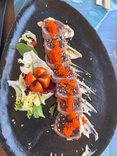A close-up, top-down shot displays a meticulously prepared dish of seared tuna and assorted salad. The tuna, sliced into thin, rare pieces, is arranged in a curved line across a black, circular plate. Each slice is lightly seared on the edges and topped with bright orange tobiko (flying fish roe) and a scattering of white and black sesame seeds. A creamy white sauce is artfully drizzled in swooping patterns behind the tuna. To the lower right, a small salad garnishes the plate, featuring frilly lettuce leaves, a few darker red leaves, and four small, quartered cherry tomatoes arranged in a flower-like pattern. A few stray sesame seeds are sprinkled across the dark plate. The background suggests an outdoor setting with a hint of a light blue, patterned placemat featuring green palm leaves, and the edge of a wooden table is visible at the bottom of the frame. The lighting suggests daytime. There is no visible text.