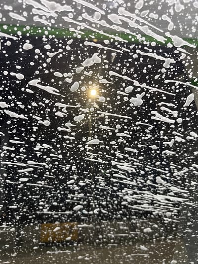 The image shows a close-up of soap suds on a dark surface, likely a window. The suds are white and form irregular patterns, with some larger streaks and many smaller droplets. A bright light source, possibly the sun, is visible through the suds, creating a glowing effect. The background is dark and out of focus, with hints of green and yellow shapes suggesting an outdoor environment. It is impossible to determine the location in the city of Berezhany, Ukraine, from this image. There are no people or discernible actions depicted.