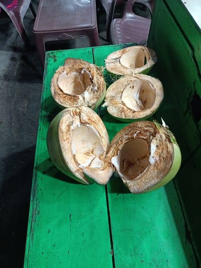 Four halved coconuts, some partially de-husked and opened, are displayed on a vibrant green wooden surface. The coconuts are in various states of preparation, with one still retaining a significant portion of its green outer layer. The interior of the coconuts reveals the white flesh and hollow cavity where the water once was. The green surface appears to be a tabletop or counter, possibly in an outdoor setting. In the background, out of focus, are hints of plastic chairs and tables, suggesting a food stall or roadside eatery. The lighting in the scene is dim, casting shadows and highlighting the textures of the coconuts and the wooden surface. The overall impression is a snapshot of a local vendor's display in Bireuen, Indonesia, likely offering fresh coconut water or produce.