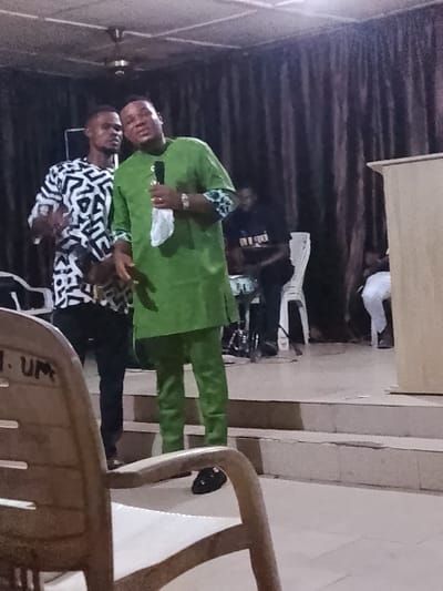 Two men stand on a stage, one in a green suit holding a microphone. The other wears a black and white patterned shirt and black pants. Behind them, a man plays drums. A person is seated on a chair to the far right. The scene is illuminated by overhead lighting, with dark curtains in the background. A wooden pulpit stands to the right. A chair is partially visible in the foreground.