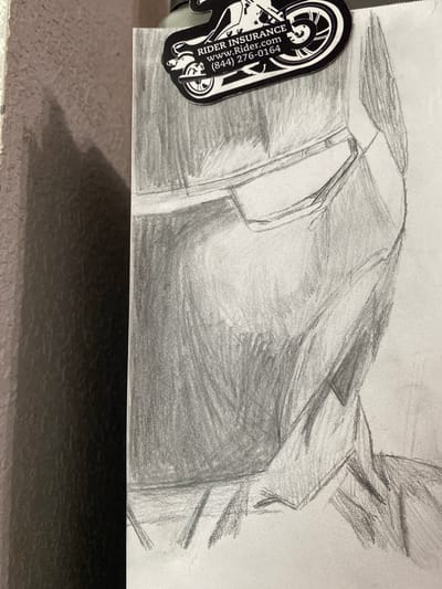 This is a pencil sketch of a helmet. The helmet is drawn in profile, with the visor and side visible. The sketch is rendered with varying degrees of shading to create depth and texture. At the top of the sketch, a sticker is attached, featuring a motorcycle wheel and text that reads "RIDER INSURANCE" and "www.Rider.com (844) 276-0164." The background of the sketch is a textured surface, possibly a wall, with a shadow cast to the left. The image appears to be a close-up, likely taken in Elizabeth City, United States, given the provided location context.