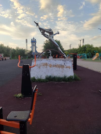 The image depicts an outdoor sports and recreation area in Temirtau, Kazakhstan. Dominating the foreground is a modern outdoor exercise machine with orange handlebars and a black seat. Behind it, a large, weathered concrete pedestal supports a gleaming metal sculpture of a person in a dynamic, athletic pose, possibly representing a swimmer or diver. The sculpture is highly reflective, catching the light from the sky.

In the mid-ground, the sports area is furnished with additional exercise equipment, some in green and orange, and a children's playground with slides and climbing structures is visible further in the background. A prominent blue and white tower with ornate detailing stands tall in the distance, bearing the Cyrillic text "Темпт". Several people are scattered throughout the area, some engaged in activities like riding a small motorcycle, while others appear to be observing or resting.

The time of day appears to be late afternoon or early evening, with soft, diffused sunlight illuminating the scene and casting gentle shadows. The sky is a mixture of blue and white clouds, suggesting fair weather. The overall atmosphere is one of active community use and leisure, with a blend of modern recreational facilities and artistic public installations. The location is clearly an urban park or public square designed for physical activity and social gathering.