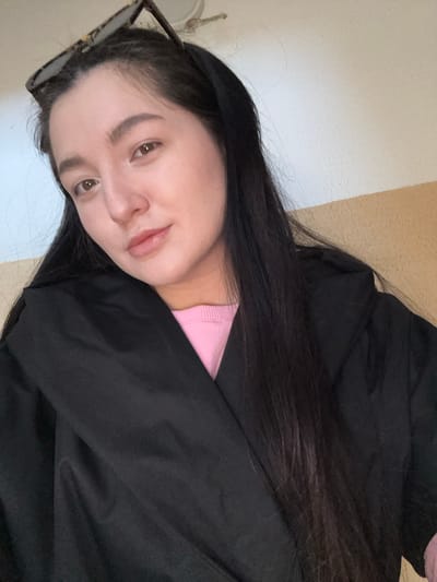 A woman with long, dark hair is lying down and looking at the camera. She has light skin, brown eyes, and full lips. She is wearing a pink shirt and a black jacket. Glasses are resting on her head. The background is a light beige wall with a white trim. The location in the city of Kurovskoye, Russia, is unknown.