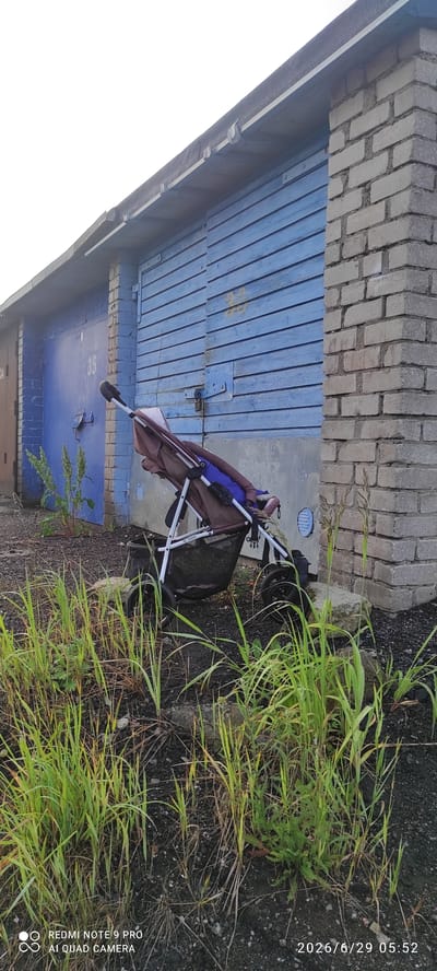 A brown and blue stroller is parked in front of a row of garages in Maardu, Estonia. The garages have blue wooden doors and one of them is marked with the number "35". There is tall green grass growing in front of the garages, and a brick wall is visible on the right side of the image. The sky is overcast. The timestamp on the image indicates it was taken on June 29, 2026, at 5:52 AM.