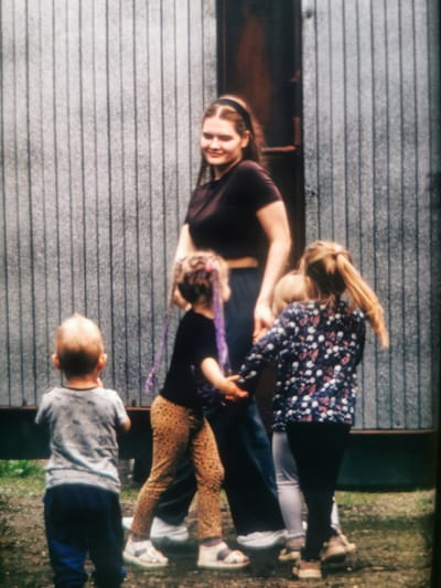 A group of five people, one adult woman and four children, stand in front of a corrugated metal wall. The woman, in the center of the frame, wears a dark brown t-shirt and dark pants. She is looking downwards and to her right, smiling. Her hair is dark brown and pulled back with a headband. To her left, a young boy with short, light brown hair faces away from the camera, wearing a grey t-shirt with a subtle pattern and blue pants. To the woman's right, a young girl with blonde hair tied in pigtails wears a black t-shirt and leopard-print pants. She is holding the woman's hand and looking down. Next to her is another child wearing a patterned shirt with a dark background and white and pink floral designs. Another child, mostly obscured, stands between the two older girls. The ground is covered with gravel and patches of grass. The corrugated metal wall is grey and appears to be part of an outdoor structure, possibly a shed or fence. This scene is taking place outside in what appears to be a residential or communal area. The location is Serov, Russia.