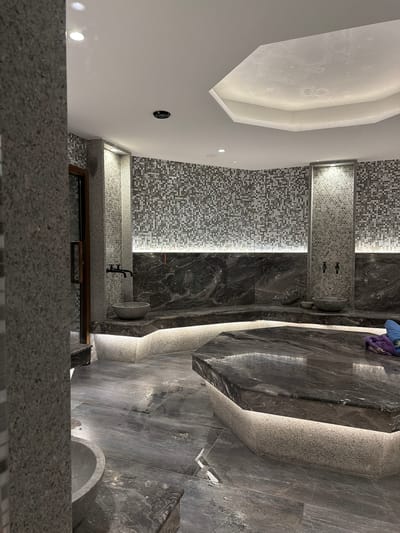 The image depicts a modern, luxurious spa or bathroom interior, likely in Hisarya, Bulgaria. The scene is dominated by dark, polished marble tiles and textured mosaic tiles, creating a sophisticated and spa-like atmosphere. A large, grey stone basin is mounted on a tiled wall, with two black faucets above it, suggesting a washing or bathing area.
In the background, there are two shower heads, also mounted on a mosaic-tiled wall. The lighting is subtle and integrated, with LED strips illuminating the edges of the tiled walls and accentuating the architectural features. One prominent feature is a large, curved bench or seating area made of textured, light-colored stone, which is softly lit from below.
The ceiling features a unique, angular design with recessed lighting, adding to the contemporary aesthetic. Part of a wooden-framed window or doorway is visible in the lower portion of the image, hinting at the transition to another area. A few folded towels, one purple and one blue, are casually placed on the edge of a marbled surface, adding a touch of human presence.
The overall impression is one of tranquility and high-end design, suitable for a spa, hotel, or private residence focused on relaxation and well-being. The time of day cannot be determined, and there is no indication of weather. No text is visible in the image.