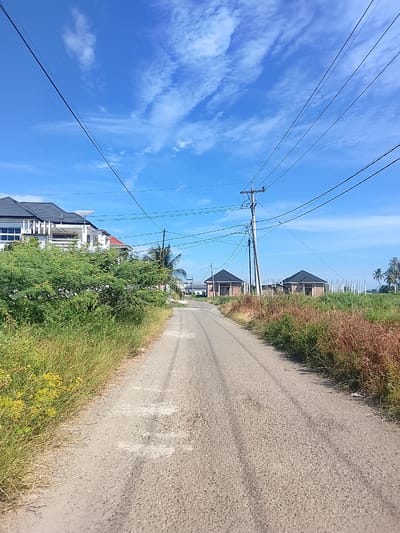 This is a photograph of a road in Rhieng Mancang, Indonesia. The road is unpaved and appears to be made of gravel. On the left side of the road, there is a dense green bush with tall dry grass in front of it. Further down the road on the left, a large white house with a dark roof is visible. On the right side of the road, there are several small houses with dark roofs and fields of dry grass. Telephone poles and power lines are visible throughout the scene, with a bright blue sky with scattered clouds overhead. The image appears to have been taken during the day, likely in the morning or afternoon, given the clear sky and sunlight.