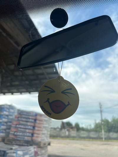 The image shows a close-up view of a car's interior looking out through the windshield. A yellow, circular air freshener with a smiley face design is hanging from a string. The smiley face has closed eyes and a smiling mouth with red lips. The word "areon" is printed in black text on the lower part of the air freshener. A rearview mirror is visible above the air freshener. The background through the windshield shows a cloudy blue sky, some trees, a building, and a stack of what appear to be bagged materials, possibly construction supplies. The location is Yizhivtsi, Ukraine.