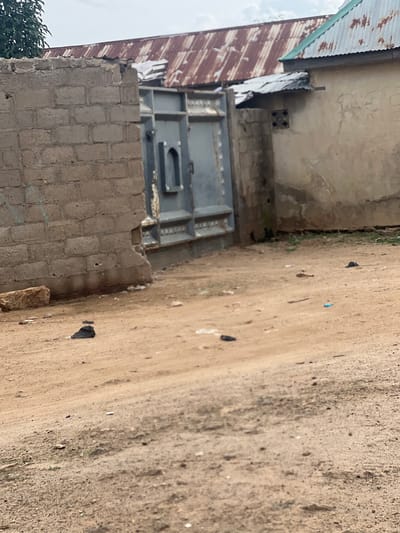 The image depicts a dusty, outdoor scene in the city of Bauchi, Nigeria. In the foreground, a sandy, unpaved ground is visible with scattered debris including small stones and some litter. A dark-colored object, possibly a piece of cloth or plastic, lies in the middle distance.
Further back, a section of a weathered, light brown brick wall with visible mortar lines stands on the left. Adjacent to it, a large, gray metal gate with intricate paneling is partially open, revealing a dark opening behind it. This gate appears to be part of a property enclosure.
To the right of the gate, a light-colored building with a textured, possibly stucco or plaster, facade is present. The building has a simple, rough finish. Above the building and the gate, a section of a reddish-brown, corrugated metal roof is visible, showing signs of rust and age. A smaller, darker section of corrugated metal roofing is also visible, extending from a lower part of the building. A glimpse of a green metal roof section is seen on the far right. The sky above is overcast with clouds. The overall impression is one of a functional, perhaps residential or commercial, area in a developing urban environment, characterized by its utilitarian structures and unpaved surfaces.
