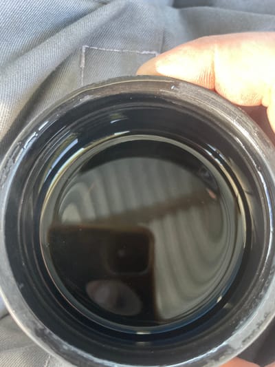 A close-up, overhead shot shows a black container filled with a dark, oily liquid. The container has ridged sides and a wide opening. The liquid appears viscous and reflects light, creating a swirling pattern on its surface. A person's hand, with visible fingerprints, rests on the rim of the container. The background consists of a textured, grey fabric, possibly clothing or upholstery. There are no visible signs of the city of Сокуры, Russia in the image.