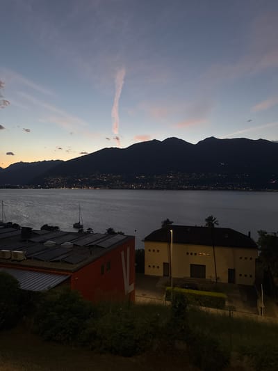 At dusk, the image captures a view overlooking Lake Maggiore from Magadino, Switzerland. The sky displays a gradient of colors from light blue at the top to a soft pink and orange near the horizon, with wispy clouds. A prominent contrail stretches vertically in the upper central portion of the sky.
In the foreground, a red building with solar panels on its roof is visible on the left. To its right, a pale yellow building with multiple windows and a dark tiled roof is situated on a slight incline. A street lamp illuminates the area in front of the yellow building.
The middle ground features the calm waters of Lake Maggiore, reflecting the fading light. Two sailboats are moored on the lake, with one appearing to be a sloop.
The background is dominated by a dark silhouette of mountains that form a ridge behind the lake. Along the base of the mountains, a city or town is illuminated by numerous lights, suggesting a settlement nestled by the water. The overall scene evokes a peaceful, twilight atmosphere in a lakeside town.