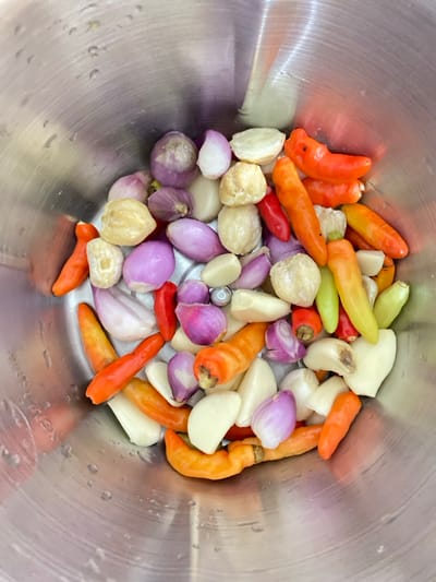 A stainless steel bowl sits on a countertop, filled with a variety of raw ingredients including red and green chilies, cloves of garlic, small red onions, and beige-colored mace. The bowl is located in Bekasi Regency, Indonesia, at coordinates -6.353 latitude, 107.193 longitude.  The ingredients are likely destined for use in a local recipe.  The scene is captured from a top-down perspective, highlighting the vibrant colors of the ingredients. The bowl is the only element in the photo.