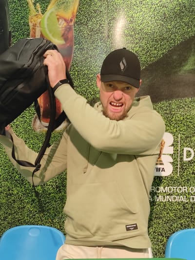A man wearing a light green hoodie and a black baseball cap with a white logo is holding a black backpack and making a grimacing facial expression with his teeth bared. He is standing in front of a green textured wall that appears to be a backdrop with images of grass, a drink with a lime wedge, and the FIFA World Cup trophy. Text on the backdrop is partially visible, including "FIFA" and "ROMOTOR O A MUNDIAL D". Two blue plastic chairs are visible in the foreground. The location is identified as Ezeiza, Argentina.