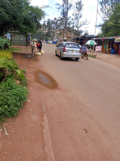 The image depicts a street scene in Murang'a, Kenya, under a partially cloudy daytime sky. The unpaved, reddish-brown dirt road runs from the foreground towards the background, curving slightly to the left. A small, irregular puddle of water is present on the left side of the road.

On the left side of the road, a dense strip of green vegetation, including bushes and a large tree, lines the foreground and midground. Behind this foliage, a green corrugated metal structure is partially visible. Further back on the left, a group of three individuals is present: one male subject in a light-colored shirt stands next to two other subjects, one in a red shirt and another in dark clothing, who are near two large, light-colored sacks on the ground beside the road. Another dark-clothed individual is partially visible further left, obscured by foliage.

In the midground, a silver Toyota Sienta (license plate KBD 659V) is traveling away from the viewer on the road. Following it is a male subject on a brown motorcycle, wearing a blue patterned shirt.

On the right side of the road, a series of brightly colored, simple commercial structures are visible. A white sign with red lettering prominently displays "BUTCHERY," and another sign to its left partially reads "CHEFFN [unclear] BUTCHERY." An open olive-green umbrella provides shade in front of one shop. Individuals are present near these shops, including one seated under a blue cover. Tall, sparse trees line the background, and buildings are visible on distant hills.