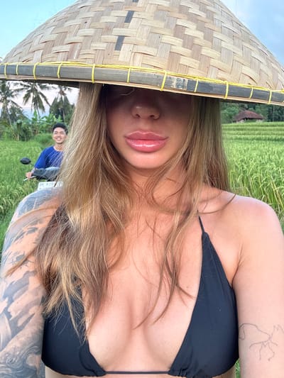 A woman is lying on her side in a rice field in Padangbulia, Indonesia. She is wearing a black bikini top and a traditional Asian conical hat. Her hair is blonde, and she has tattoos on her arms. A man is sitting on a scooter behind her, smiling. The rice field is green and lush, and the sky is cloudy.