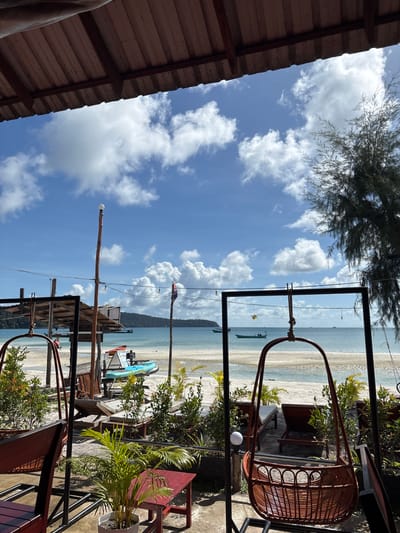 The image captures a view from a covered outdoor seating area looking out towards a tropical beach and the ocean. In the foreground, two hanging rattan swings are visible, with one partially obscuring a small red table and a potted palm plant. Behind the swings, a sandy beach stretches out, dotted with lounge chairs and more greenery. Further in the distance, calm, turquoise water meets a hazy, tree-covered island under a bright blue sky with scattered white clouds. Several small boats are anchored or moving on the water. A rustic wooden structure with a corrugated metal roof stands on the left, and flagpoles with flags are visible. The scene suggests a relaxed beachfront establishment in Preah Sihanouk, Cambodia, likely a cafe or restaurant.