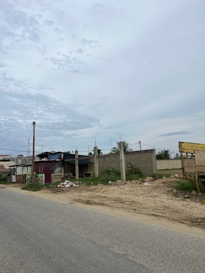 A roadside scene in Landuh, Indonesia, shows a mixture of structures and debris under an overcast sky. A paved road occupies the foreground, with a dirt shoulder bordering it. The shoulder is strewn with trash, including plastic bottles and paper.
Several small, makeshift buildings are visible on the left side of the road. One appears to be a small shop with colorful posters advertising drinks. Adjacent to it are structures with corrugated metal walls and tarpaulin roofs, some in disrepair. A wooden pole with electrical wires stands prominently near these buildings.
Further into the scene, a low, grey brick wall runs horizontally. In front of the wall, several concrete pillars stand at intervals, some with rebar protruding from the top, suggesting ongoing or incomplete construction. Behind the wall, more buildings and greenery are visible, including palm trees. On the far right, part of a weathered yellow truck or trailer is visible, its wooden structure exposed. The ground is uneven, with patches of dry grass and exposed soil.