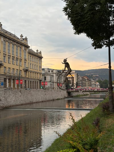 A sculpture of a cyclist in motion is the central subject, suspended in mid-air above a street. The cyclist is crafted from metal, with intricate detailing suggesting movement and effort. Behind the sculpture, buildings line the street, with a mix of architectural styles visible, some appearing older and ornate, others more modern. A canal or river runs alongside the street, its surface rippling and reflecting the warm tones of the sky. Greenery, including trees and bushes, frames the scene, adding a touch of nature to the urban environment.
The time of day appears to be either sunrise or sunset, given the soft, golden light illuminating the sky and reflecting in the water. The sky itself is a blend of soft blues and oranges, with clouds present. The weather seems calm and pleasant.
In the background, a hilly landscape is visible, dotted with more buildings, suggesting the location is nestled within a valley or at the edge of a city. The scene is tranquil, with a sense of stillness, broken only by the implied movement of the cyclist sculpture.
There are several pieces of visible text on red banners attached to lampposts along the street. Though small and somewhat blurry, they appear to contain the word "MARKET" or a similar word in English, repeated. A green traffic light is also visible in the distance. The overall impression is one of a peaceful urban setting during the twilight hours, enhanced by the artistic presence of the cyclist sculpture and the natural beauty of the water and sky.
