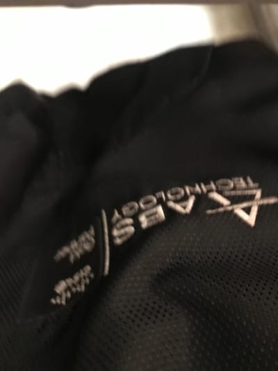 The image displays a severely out-of-focus, close-up view of black fabric with a white label or embroidery. The fabric exhibits a fine, perforated or mesh texture in areas. The legible text on the label, oriented vertically and largely upside down, includes "AXLABS TECHNOLOGY" and "ABS". Additional smaller, white text is visible but illegible. The upper portion of the frame shows a blurred white area, likely an overhead light source or a bright surface. There are no individuals, discernible actions, or environmental details present to determine a specific location within Calabar, Nigeria. The scene is static, focused solely on the textile detail.