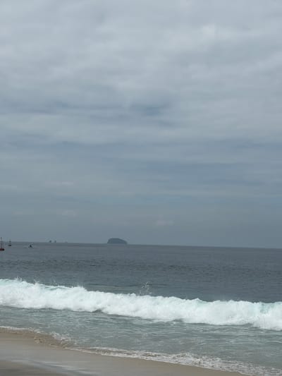 The image depicts a serene beach scene in Manggis, Indonesia. The ocean is calm with small waves gently lapping the sandy shore. In the distance, a few islands are visible, shrouded in a light mist. The sky is overcast with grey clouds, casting a soft light on the scene. There are no people visible in the image, nor any signs of activity. The overall impression is one of peace and tranquility, with the natural beauty of the Indonesian coastline on full display.