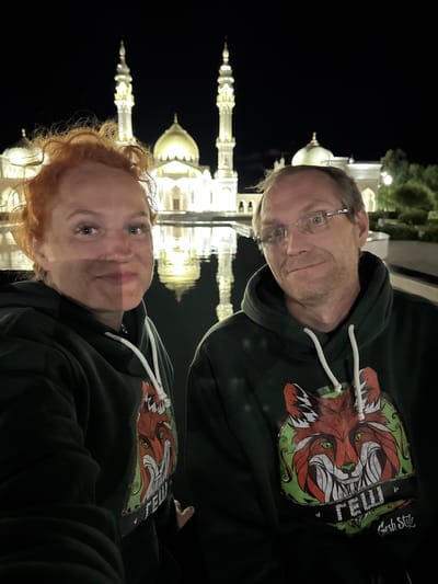 This is a nighttime, outdoor, eye-level, selfie-style photograph taken in the city of Bolgar, Russia. In the foreground, two individuals, a woman and a man, are positioned close to the camera, both wearing dark green hooded sweatshirts with a graphic of a fox's face and text below. The woman, on the left, has red curly hair and is smiling. The man, on the right, wears glasses and has short brown hair and stubble. Behind them, a white mosque with multiple domes and minarets is illuminated by lights. The mosque is reflected in a body of water in front of it. Trees and other foliage are visible in the background.