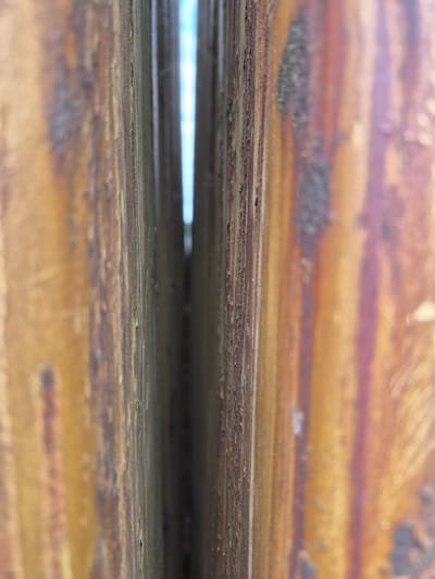 This is a close-up, macro photograph of a narrow gap between two weathered, vertical, metallic structures. The surfaces of these structures are heavily textured and show significant signs of rust, with streaks of brown, orange, and yellow dominating the color palette. The textures appear rough and degraded, with peeling paint or material visible along the edges. A sliver of bright, possibly sky-blue light is visible through the gap, suggesting the structures are outdoors. The photograph is taken from a low angle, emphasizing the verticality of the structures and the depth of the gap. There are no people or discernible actions captured in the image. The context of Masów, Poland, is not visually represented in the provided image itself.