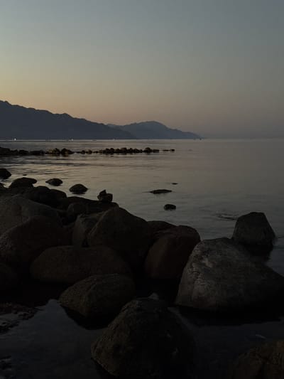 A low-angle, wide shot captures a tranquil seascape at dusk, likely in Dahab, Egypt, given the characteristic mountainous terrain bordering the Red Sea. The foreground is dominated by dark, wet rocks of varying sizes scattered along the shore. Some rocks are partially submerged in the still, dark water, reflecting the dim light. Beyond the immediate rocks, a line of smaller rocks extends into the sea, creating a subtle barrier. The water itself is calm and reflective, mirroring the muted colors of the sky. In the distance, a hazy mountain range forms the horizon, its silhouette softened by the fading light. A faint glow emanates from scattered lights along the distant shoreline, suggesting a settlement or a promenade. The sky transitions from a pale yellow near the horizon to a muted blue-grey overhead, indicating the transition from sunset to twilight. No people are clearly visible, although faint silhouettes might be discerned on the distant shore. The overall atmosphere is serene and peaceful.