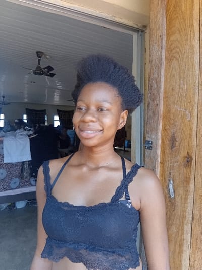 An image taken in Ejule, Nigeria, depicts a young adult female standing in an open doorway. She has dark skin, short, dark, kinky hair styled in an upright fashion, and is smiling, revealing her teeth. She wears a dark blue or black lace crop top with thin straps, beneath which a dark bra is visible. A small earring is present in her left ear. To her right, a weathered wooden doorframe exhibits vertical grain. The background reveals an interior space with a white paneled ceiling equipped with multiple dark-colored ceiling fans, at least two of which are distinct. Furniture draped with patterned fabric, a white garment, and other indistinct objects and figures are visible within the room behind her.
