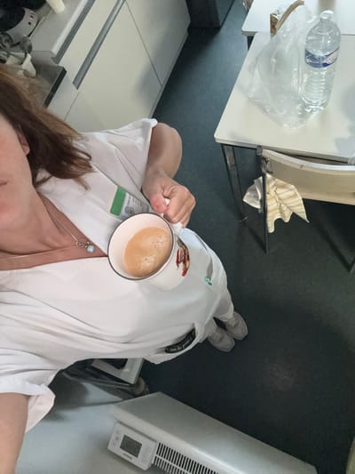 An overhead shot captures a person, likely female with brown hair, dressed in a white uniform or scrubs, holding a white mug filled with a light brown, foamy beverage, probably coffee. The person's face is mostly out of frame, but a relaxed posture is suggested as they hold the mug near their chest. A necklace with a light blue or opal-like bead pendant is visible, along with a lanyard or badge holder featuring a green section.
The setting appears to be an indoor professional environment, possibly a break room or utility area within a medical facility. The floor is dark gray or black. To the left, a white table or counter holds a clear plastic water bottle, identifiable as "CRISTALINE" with "eau de source" partially visible, resting in a clear plastic bag. Below the table, a striped towel and another white cloth are neatly folded on a lower shelf.
To the right, a white air conditioning or ventilation unit with a digital display showing "0.0" and several control buttons is mounted on the wall. Part of a light-colored counter or appliance is visible in the lower left. Light-colored sneakers are visible on the person's feet, further suggesting a practical, work-related setting.
Visible text on the green section of the lanyard includes "Centre Ho..." which likely refers to "Centre Hospitalier" (Hospital Center), and additional letters "F..." and "L...". The brand name "CRISTALINE" is clearly legible on the water bottle. On a black tag or ribbon attached to the uniform sleeve or collar, partial text "Du grand R..." can be seen. Given the location context of Tourcoing, France, the "Centre Hospitalier" detail strongly suggests a hospital environment in that city. The image captures a moment of break during work.