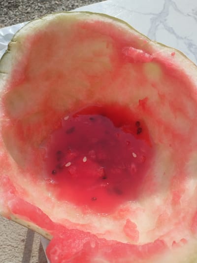 An overhead, close-up shot captures the interior of a hollowed-out watermelon rind. The rind, primarily green on the outside and white on the inside, is now filled with a pool of pink watermelon juice. Several watermelon seeds, both black and white, are scattered within the juice and on the wet inner surface of the rind. The watermelon appears to have been recently eaten, with some flesh still clinging to the rind. The background shows a textured surface, possibly a countertop or table, with a hint of a marble pattern, and a blurry granular surface beneath. The lighting suggests it is daytime. The context of Чагыр, Kyrgyzstan is not directly evident from the image itself.