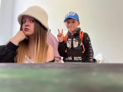 Two individuals are positioned in the lower half of the frame against a pale background. On the left is a young boy wearing a blue cap with the word "CAMP" printed on it, along with a black hoodie featuring orange straps and graphic designs. He is holding up two fingers in a peace sign. To the right of the boy is a young woman with long blonde hair and a white furry hat. She has one hand near her face. Above the subjects is a blurred, indistinct object that obscures the background. The location is unspecified and the image does not give any evidence to identify the city of Pushchino, Russia.
