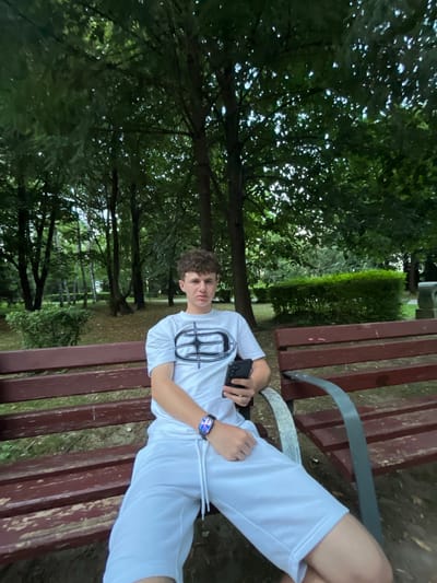The media file shows a young man sitting on a park bench. He is wearing a white t-shirt with a black graphic design and white shorts. He is holding a black smartphone in his right hand. On his left wrist, he is wearing a smartwatch displaying the time "18:49". The bench is made of dark brown wood with metal supports.
The background is a park with lush green trees and foliage. The lighting suggests it is daytime, possibly late afternoon given the time on the smartwatch. The overall mood is relaxed and casual. The location is identified as Gotse Delchev, Bulgaria. There is no discernible text in the image other than what is displayed on the smartwatch.
