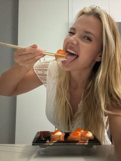 A young woman with long blonde hair is holding a piece of sushi with chopsticks, about to eat it. She is winking and sticking out her tongue, appearing playful and happy. She is wearing a white lace-trimmed top. Sushi rolls are visible in a black plastic container next to her. The setting appears to be indoors, possibly a home or restaurant, with plain walls in the background. The lighting suggests it could be daytime. The overall mood is lighthearted and fun. No visible text is present in the image.