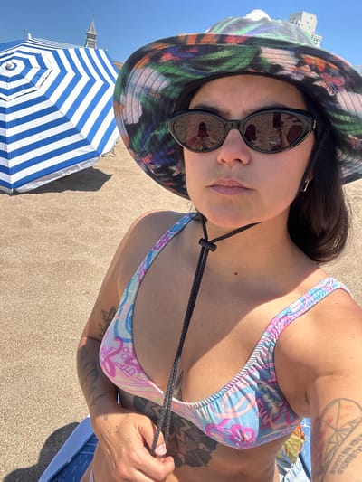 A woman wearing a floral bucket hat and sunglasses poses at the beach. She is wearing a patterned bikini top. A blue and white striped beach umbrella is visible in the foreground. Tattoos adorn her arms and chest. The beach setting features tan sand.