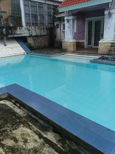 The scene depicts an outdoor swimming pool area in Pô Kô, Vietnam, with surrounding structures exhibiting signs of disrepair. A rectangular swimming pool, tiled in light blue and filled with clear, turquoise water, occupies the center and foreground. Steps descend into the water from the right side. The pool perimeter is bordered by dark blue tiles.

To the right, a two-story building with a red tiled roof features peeling purple paint on its walls, revealing lighter underlayers. The facade is marked with extensive graffiti, including the visible phrase "F*** You" in black. Two large white double doors are centered on the building's face, flanked by two decorative pillars with stone bases. Two wall-mounted light fixtures are present on either side of the doors.

To the left and behind, a multi-story concrete structure with multiple gridded windows or shutters shows significant staining, moss, and grime on its walls. A blue tiled ramp or slide descends from this structure towards the pool area.

The ground immediately surrounding the pool includes patches of bare earth, concrete, and areas covered with moss and sparse vegetation. A dark coiled rope lies on the dark-tiled edge of the pool near the entry steps on the right. No individuals are present in the image.