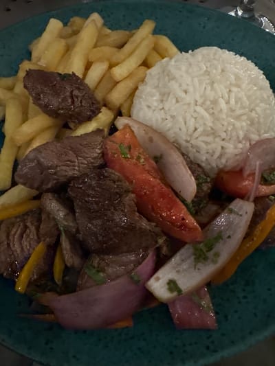 A top-down shot reveals a plate of Peruvian Lomo Saltado. A heap of white rice sits alongside a pile of french fries. Slices of sauteed beef are mixed with sliced onions, peppers, and tomatoes. The meal rests on a speckled turquoise plate. The location is likely Ancón, Peru, a city known for its coastal cuisine and the origin of the dish. No people are visible. The interaction centers around the culinary experience represented by the prepared meal.