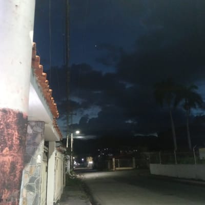 The image depicts a street scene at dusk or night in El Limón, Venezuela, with a dramatic, dark, and cloudy sky. In the foreground, a white pillar with textured, reddish-brown staining on its lower portion is visible to the left. This pillar is partially covered by stonework featuring irregularly shaped, light-colored stones outlined in white grout. A residential building with a tiled roofline in shades of orange and a light-colored overhang runs along the side of the street. The street itself is paved and empty, illuminated by a distant streetlamp. Tall utility poles with electrical wires stretch across the upper part of the frame. Two palm trees are silhouetted against the dark sky. A bright point of light, possibly a star or planet, is visible high in the sky amongst the clouds. In the background, more residential structures and fencing can be observed, with some lights indicating activity. The overall atmosphere is moody and quiet.