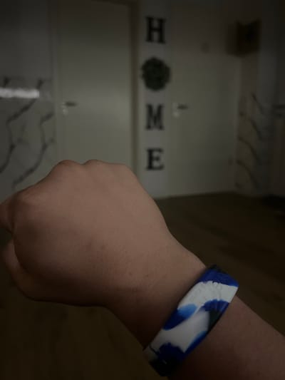 The image shows a person's arm with a blue and white patterned bracelet. In the background, the word "HOME" is spelled out in large letters on a wall. The setting appears to be indoors, possibly a home environment, with what looks like doors and patterned walls visible. The lighting is dim. There is no information in the image that places it in Bad Zwischenahn, Germany.